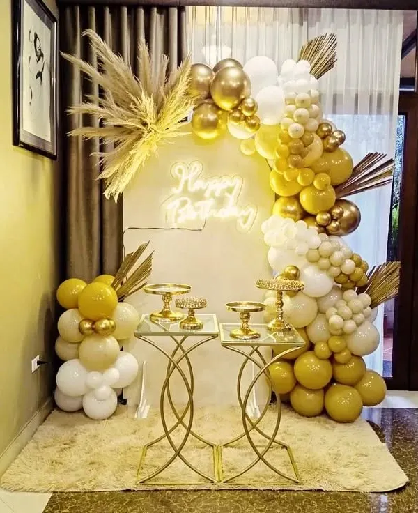 This full boho-glam setup features a half-arch balloon garland in gold and white, accented with fluffy pampas grass. The 'Happy Birthday' neon sign adds a modern touch, while the gold dessert tables provide a stylish space for the cake and treats.