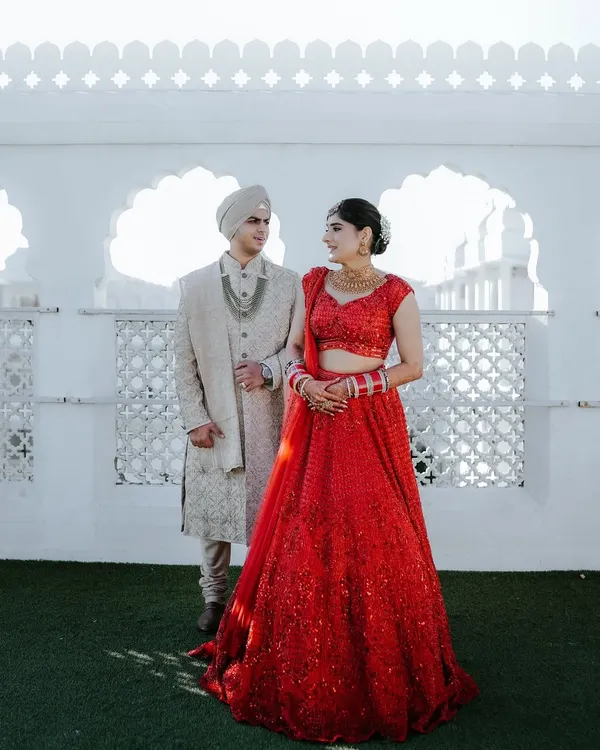 A stunning full-length portrait of the couple against a white architectural backdrop. The bride's sequined red lehenga and the groom's classic sherwani create a timeless look for their royal pre-wedding shoot.