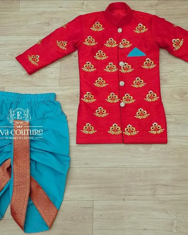 A handsome sherwani-style kurta and dhoti set for boys. The bright red jacket has intricate gold embroidery and is paired with a contrasting turquoise dhoti.