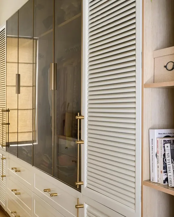 This elegant walk-in closet in our 'House of Kismish' project features a blend of white PU-finished drawers, tinted glass shutters with brass handles, and louvered panels for ventilation.