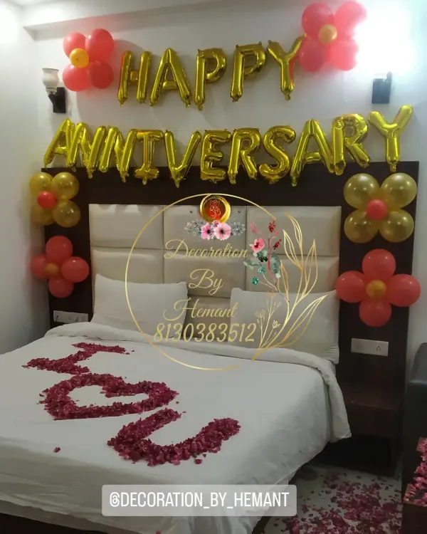A romantic anniversary decoration right in the bedroom. The setup includes "Happy Anniversary" foil balloons above the headboard, simple balloon clusters, and "I Love You" written with rose petals on the bed.
