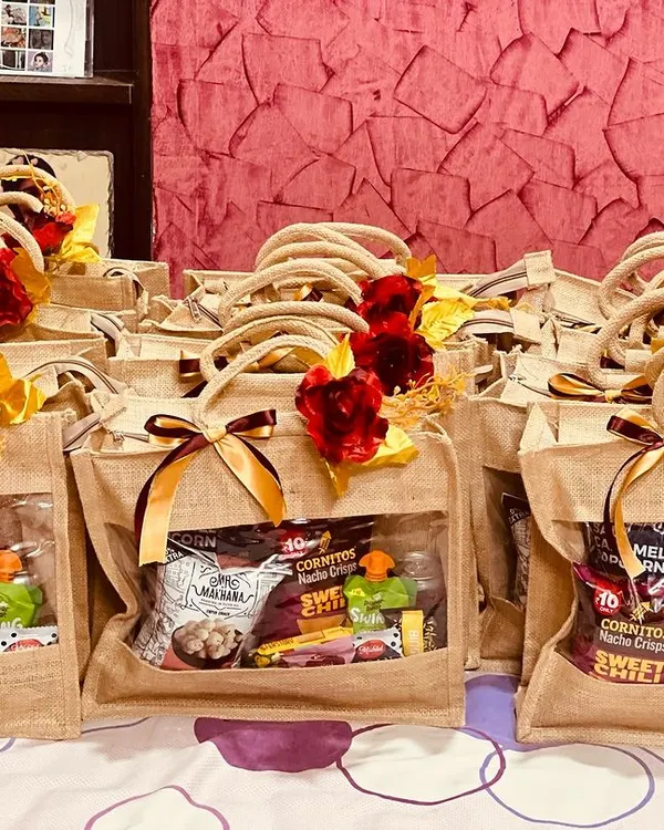 A large batch of wedding room hampers, ready to welcome guests. These eco-friendly jute bags are filled with snacks and drinks, and decorated with a red rose and ribbon.