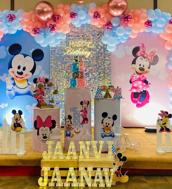 A grand Mickey and Minnie Mouse theme for a twin's first birthday. The stage is split into blue and pink sections, each with large character backdrops, a shimmer wall, and personalized props.