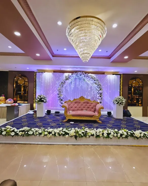 A wide view of a stage designed for a ring ceremony in a banquet hall. The setup includes a floral arch, a pink and gold sofa, and a stage skirted with white flowers.