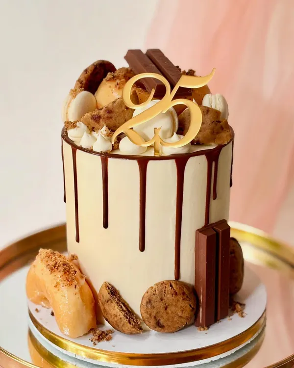 A close-up of my popular loaded drip cake for a 25th birthday. You can see the rich chocolate drip, cookies, donuts, macarons, and a custom gold topper.