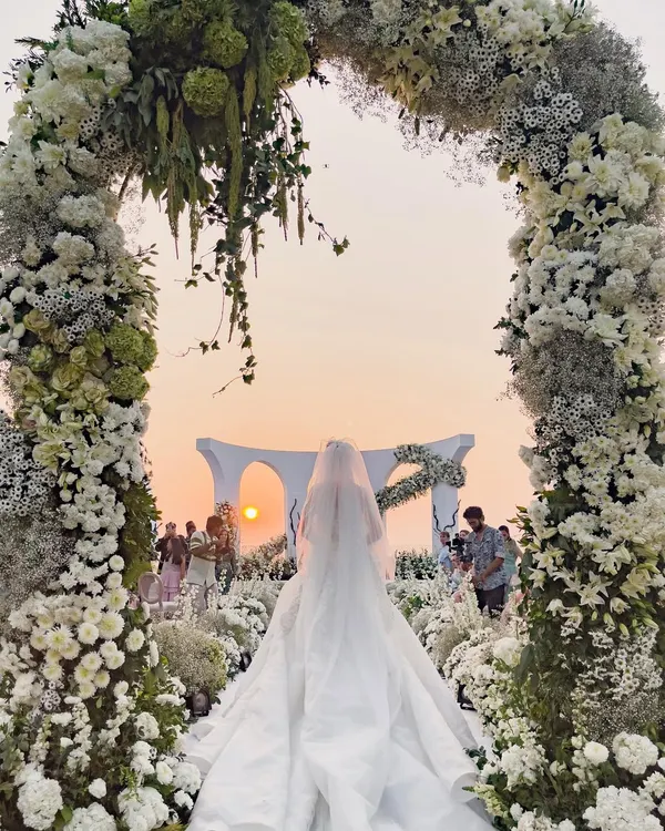 A fairytale moment as the bride walks towards a breathtaking sunset ceremony. The floral archway frames the scene, creating a perfect, romantic path for her grand entrance at this stunning white wedding in Goa.