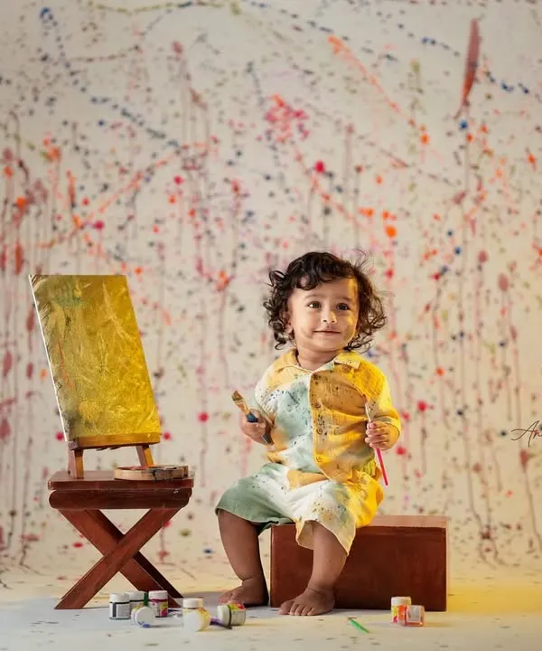 A toddler dressed as a little artist smiles proudly next to his painted canvas on an easel, against a paint-splattered wall.