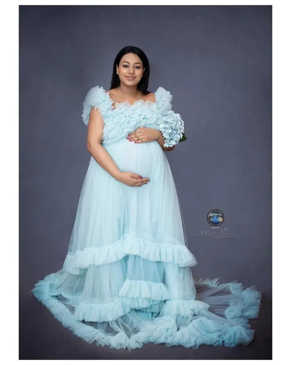 A full length portrait showcases the beauty of a flowing maternity gown. This mother to be looks radiant in a layered, light blue tulle dress, holding a simple bouquet that adds a soft, delicate touch to the image.