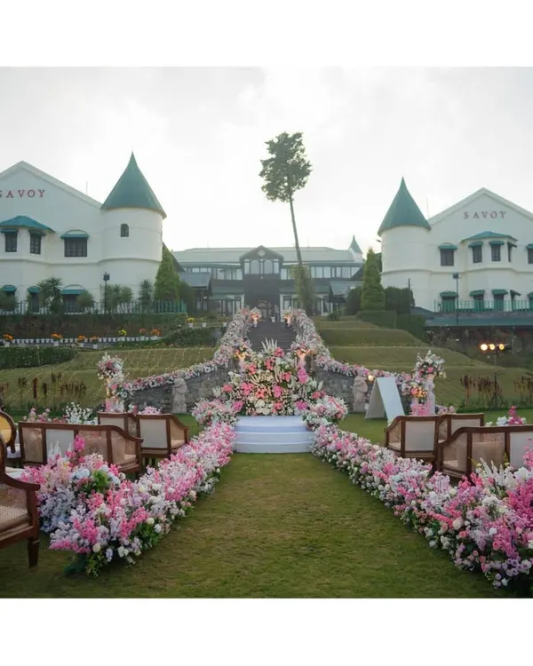 The stage is set for a fairytale wedding in the hills of Mussoorie. The aisle, lined with lush pink and white florals, leads to a mandap with the majestic Savoy palace as its backdrop, promising a ceremony of unparalleled beauty.