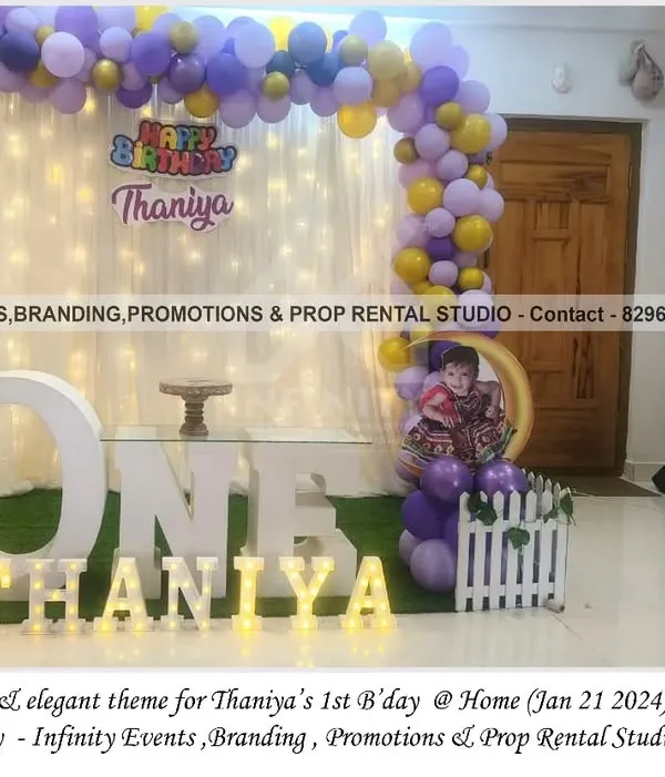 A simple and elegant theme for Thaniya's first birthday at home. This setup features a white curtain backdrop with fairy lights, a purple and gold balloon garland, and a "ONE" table, creating a magical corner.