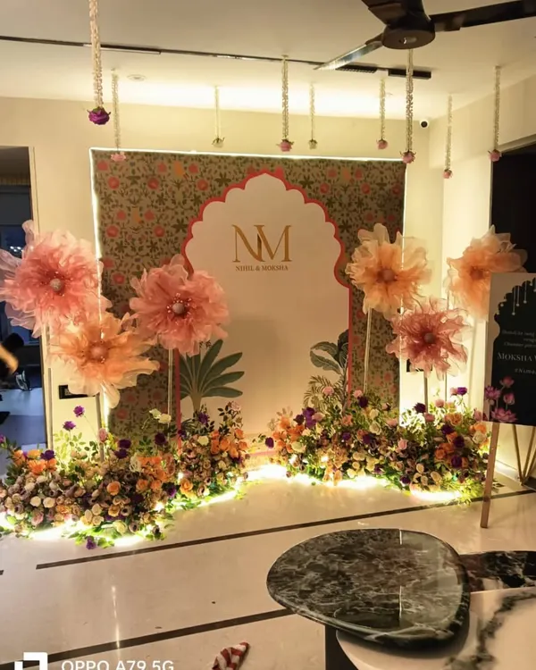 An elegant backdrop for an engagement or reception, featuring a custom monogram (NM) in a Mughal-style arch. The setup is decorated with large handmade paper flowers and fresh floral arrangements.