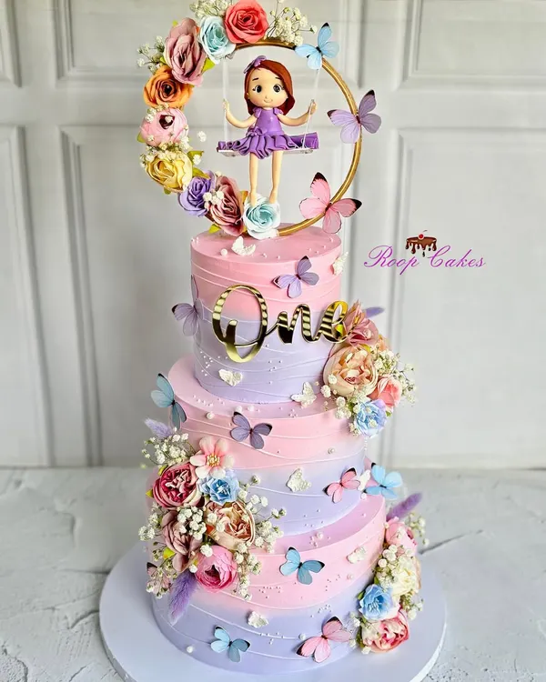 A whimsical and dreamy three-tier cake for a magical first birthday. This pastel creation is adorned with delicate butterflies, blooming florals, and an adorable fondant girl on a swing.