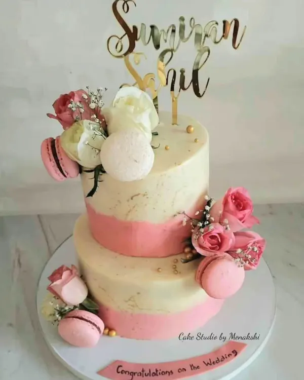 A beautiful two-tier wedding cake in white and pink. This no-fondant design is finished with buttercream and adorned with fresh roses and delicate macarons for a touch of elegance.