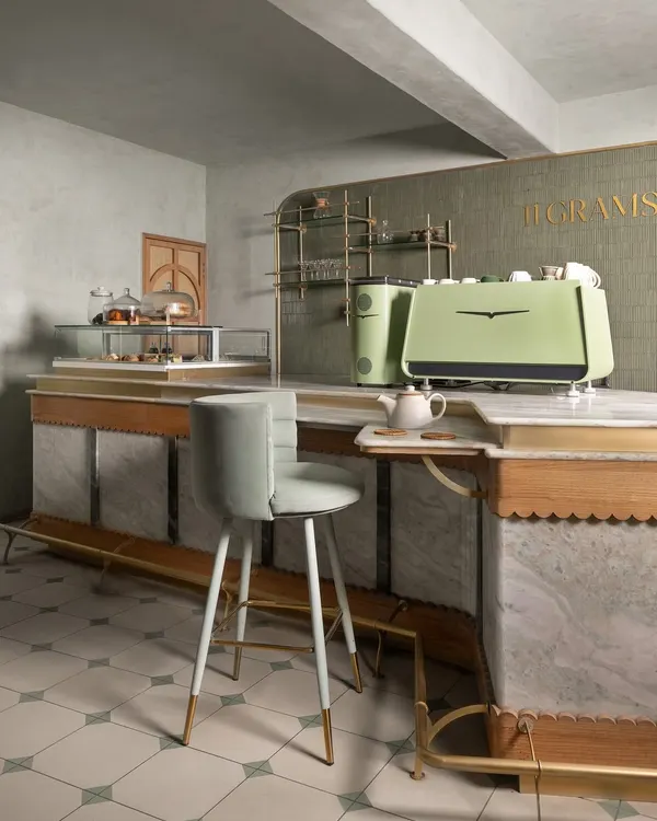 The main service counter at 11 Grams is crafted from white marble with delicate scalloped wood trim and brass details. The design ensures operational flow while serving as a beautiful centerpiece, complemented by a custom pale green espresso machine.