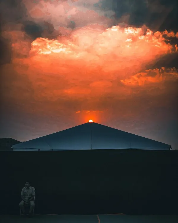 A minimalist and symmetrical composition of the sun setting behind a large tent, with a lone security guard seated, edited with a dramatic orange and black contrast.