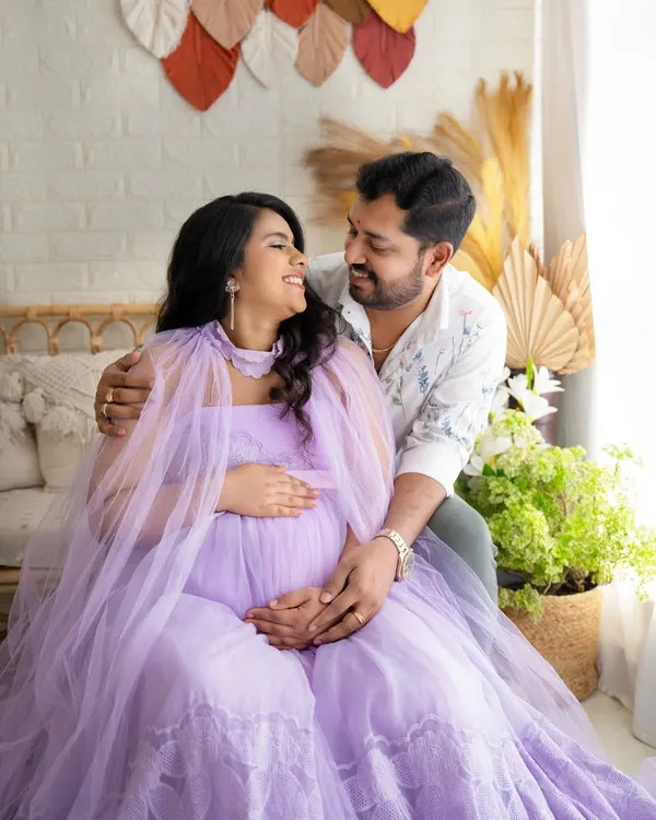A happy, interactive moment between an expecting couple. The mother-to-be, in a lovely lavender gown, shares a smile with her partner in a bright, boho-chic studio setting.