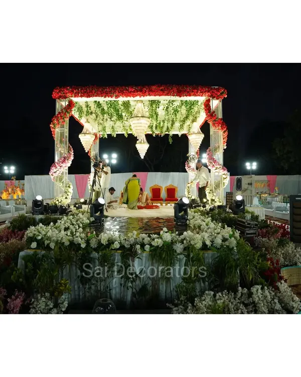This is a large, open-air wedding mandap I set up for a night ceremony. The structure is decorated with red and white flowers, hanging chandeliers, and is surrounded by a garden of white flowers for a truly grand celebration.