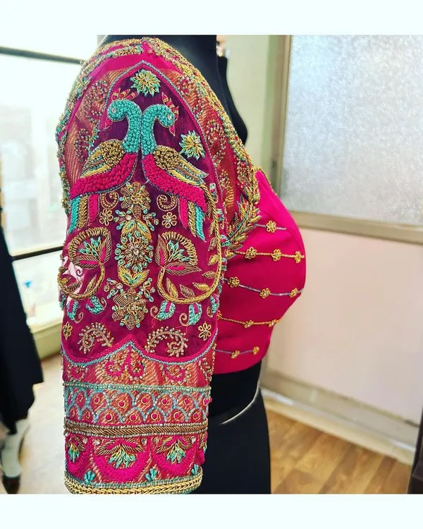 A side view of a heavily embroidered bridal blouse sleeve. The design features paired peacock motifs in zardosi and threadwork, creating a rich and traditional look for a South Indian bride.