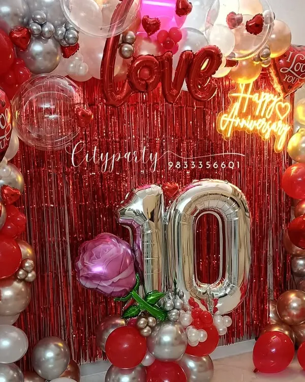 A complete view of our 10th-anniversary balloon decor package. It includes a full balloon arch, a red tinsel backdrop, large '10' number balloons, and a 'Happy Anniversary' neon sign.