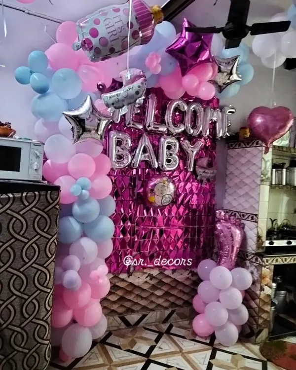This is a popular and affordable welcome baby setup for your home. It includes a bright pink foil curtain, silver 'WELCOME BABY' balloons, and balloon pillars in a mix of pink, blue, and purple.