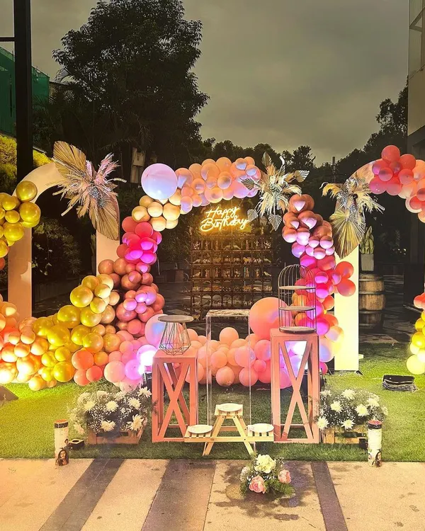 A glamorous outdoor birthday setup under the evening sky. This arrangement features twin circular balloon arches in warm sunset tones, a central neon "Happy Birthday" sign, and elegant pedestal tables, creating a sophisticated and inviting party atmosphere.
