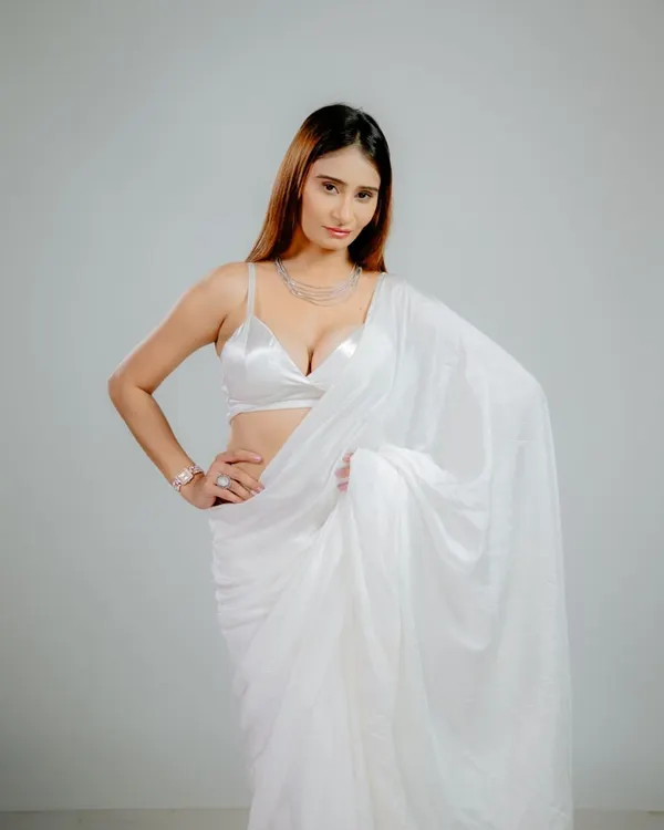 A full-length portrait against a clean grey background. The bright lighting beautifully illuminates the white satin saree, highlighting its texture and drape for a glamorous fashion shot.