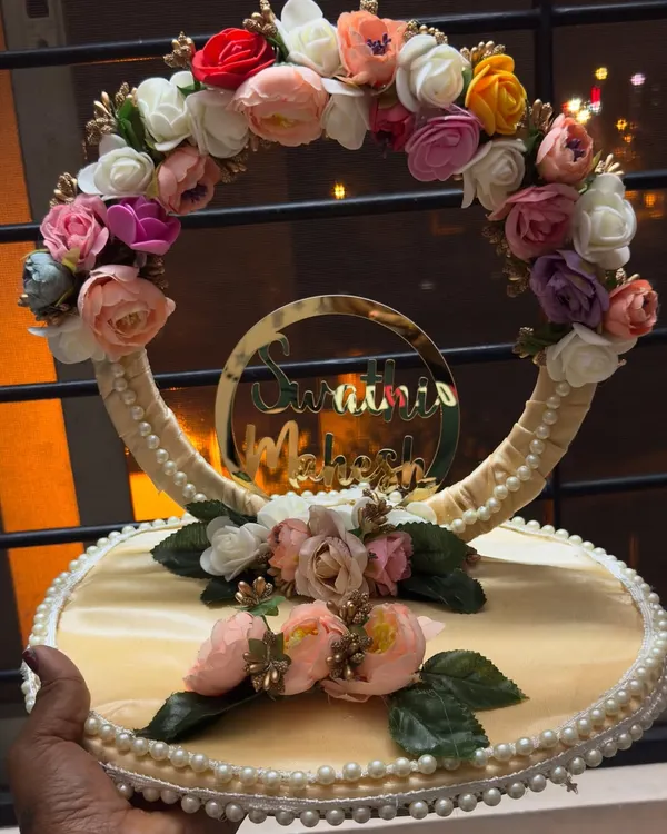 A close-up of a customized engagement ring platter. This design features a floral wreath, pearl detailing, and a golden acrylic cutout of the couple's names, Swathi and Mahesh.