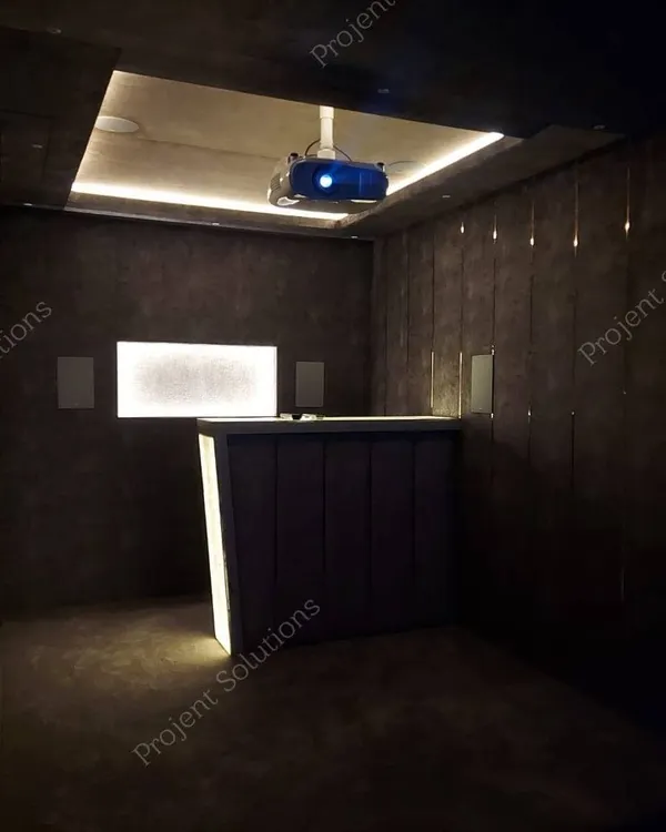 An ambiently lit view of a theater's rear section, highlighting the custom bar, wall-mounted speakers, and the glow from the ceiling-mounted projector.
