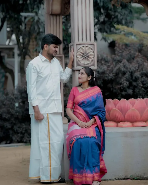 A beautiful pre-wedding portrait of a couple in traditional attire. The bride's vibrant silk saree and the groom's classic dhoti are perfectly complemented by the elegant stone pillar.