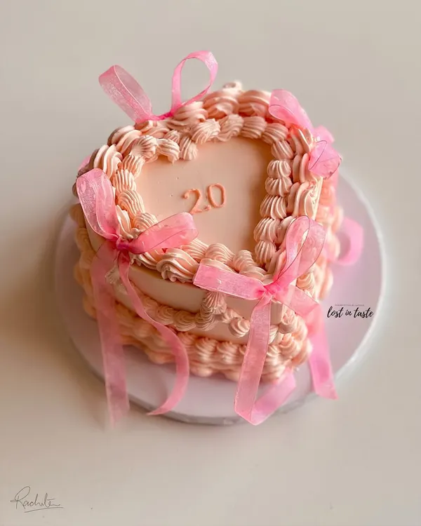 A beautiful heart-shaped cake in a soft pink hue, decorated with classic vintage Lambeth piping and delicate organza ribbons. This design is perfect for celebrating a 20th birthday in style.