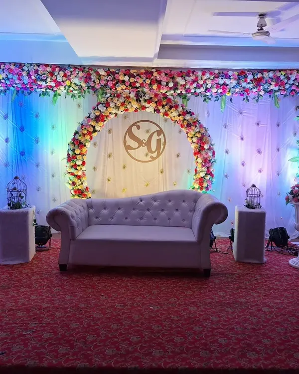 A complete stage view for an engagement, featuring a plush sofa for the couple. The backdrop includes a semi-circular floral arch and is lit with colorful spotlights to create a festive and romantic setting.