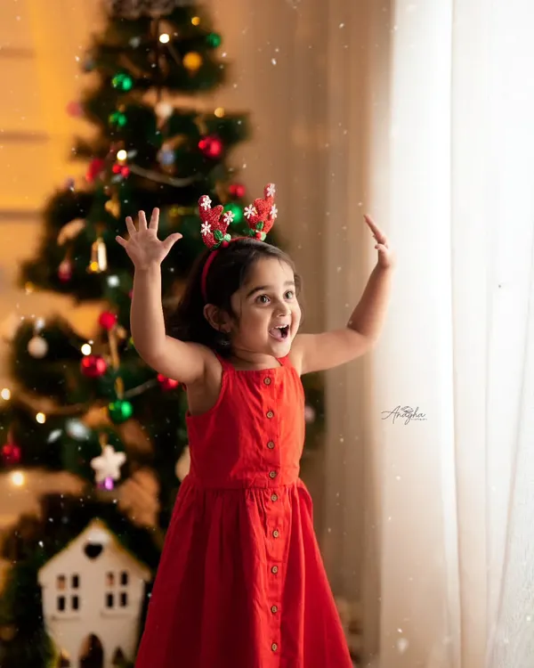 A young girl in a red dress and reindeer antlers raises her hands in excitement in front of a decorated Christmas tree.