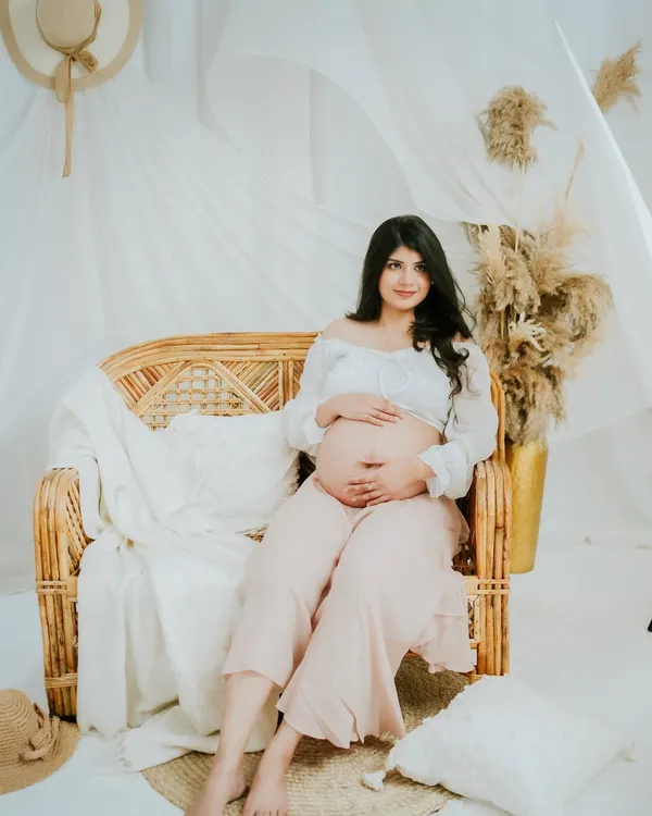 A full-length portrait from a boho-themed maternity session. The rattan furniture, pampas grass, and soft fabrics create a warm, earthy, and relaxed atmosphere.