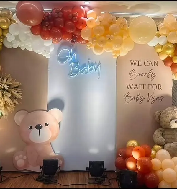 This teddy bear baby shower decor combines a warm color palette of red, gold, and peach with an "Oh Baby" neon sign. The setup includes two cute bear props and is personalized with the baby's name on the backdrop.