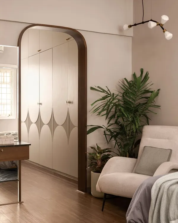 This bedroom corner is a study in calm. A custom wardrobe with a subtle geometric pattern is framed by a dark wood arch, while a comfortable armchair and a large plant create a perfect spot for relaxation.