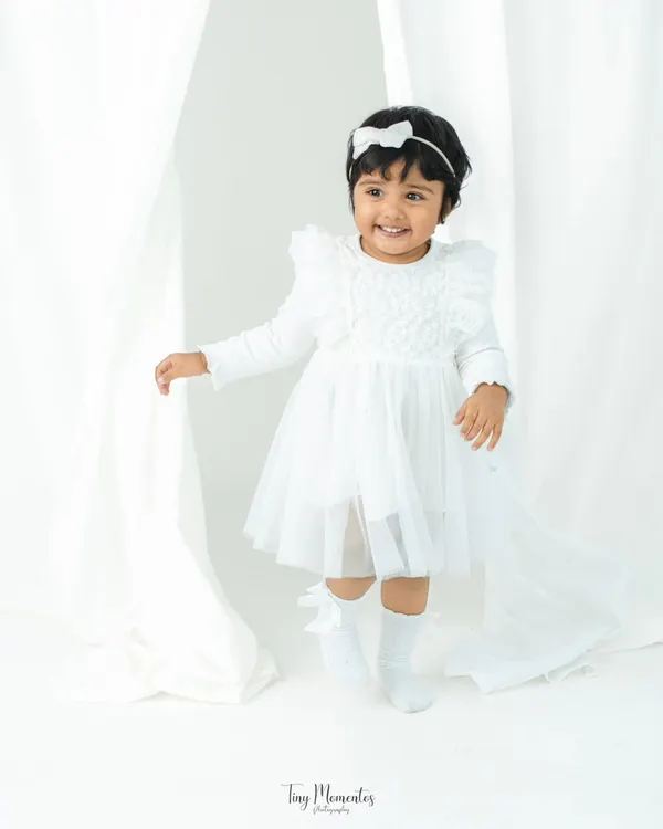 A vision in white. This two-year-old girl's bright smile and playful energy are captured beautifully against a simple, elegant white curtain backdrop.