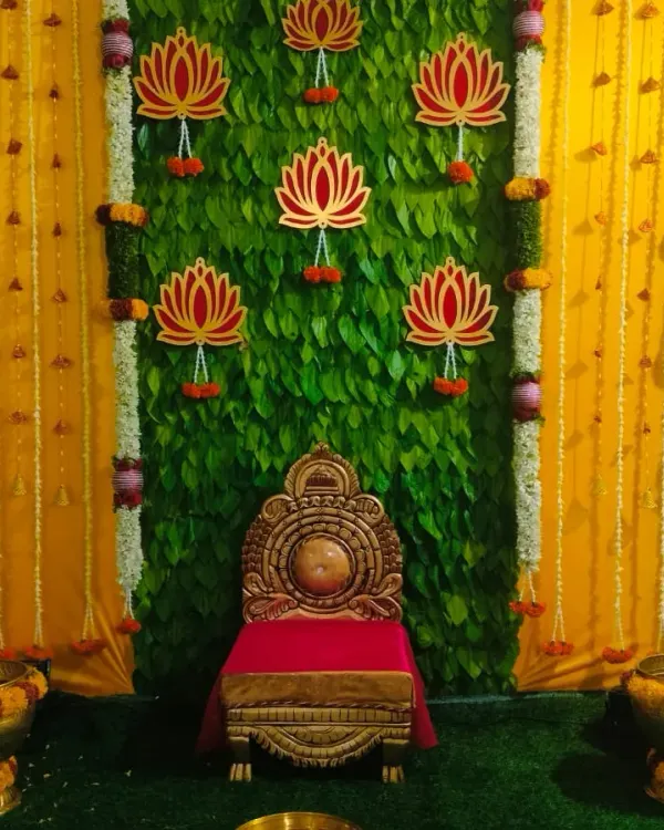 A beautiful setup for a half saree function. The backdrop is a wall of fresh betel leaves, accented with lotus cutouts and framed by yellow drapes and flower columns.