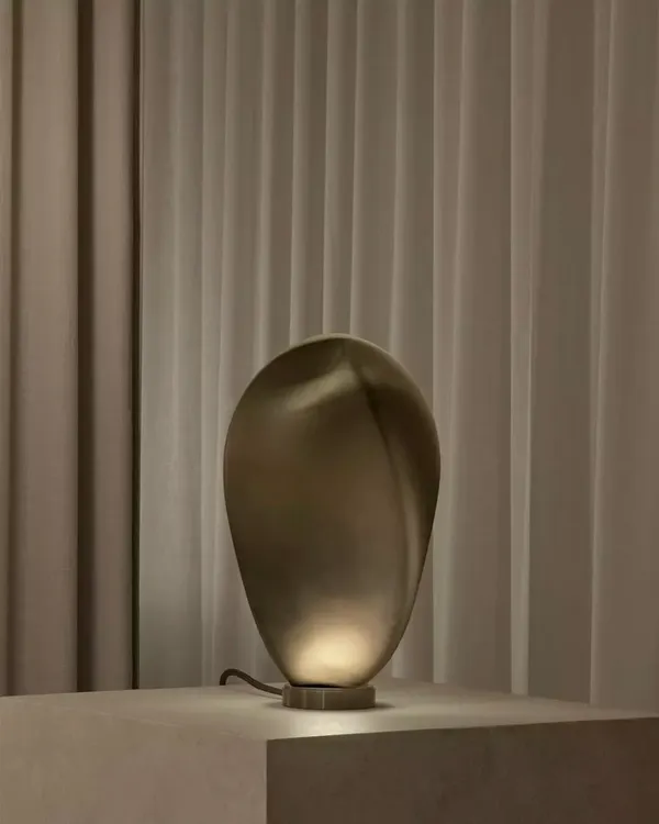 The Solace table lamp by Ross Gardam, with its visually enchanting teardrop shape, sits on a pedestal against a soft curtain. The bronze glass finish creates a warm, moody light, making it a perfect accent piece for a sophisticated interior.
