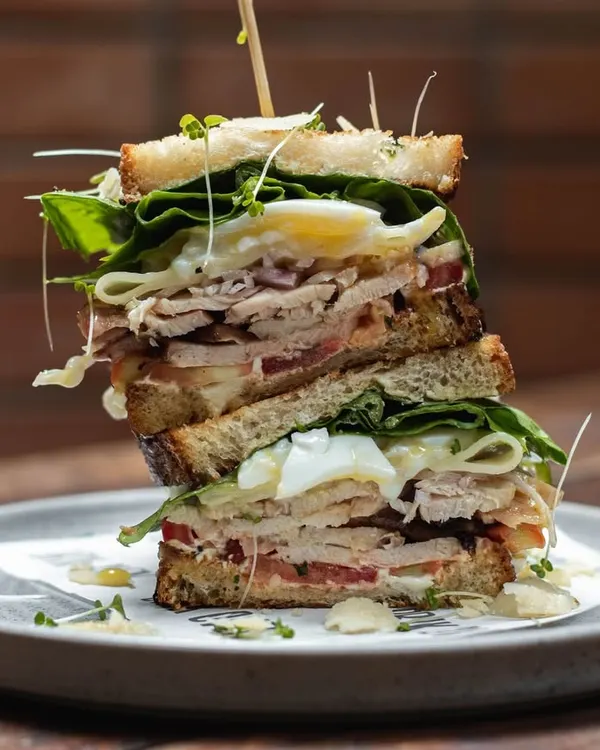 A towering club sandwich, stacked high with roasted chicken, bacon, egg, and fresh greens between slices of our toasted sourdough bread. This is a proper meal.