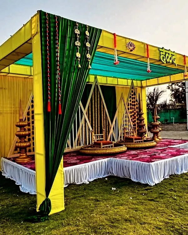 A full view of a beautiful outdoor Haldi mandap setup. The structure is decorated with yellow and green drapes, traditional brass lamps (diya stands), and comfortable seating for the main ceremony.