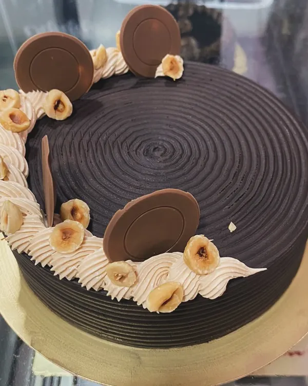 A rich chocolate hazelnut cake. This design features a textured chocolate ganache, hazelnut buttercream, and is topped with toasted hazelnuts and chocolate discs.