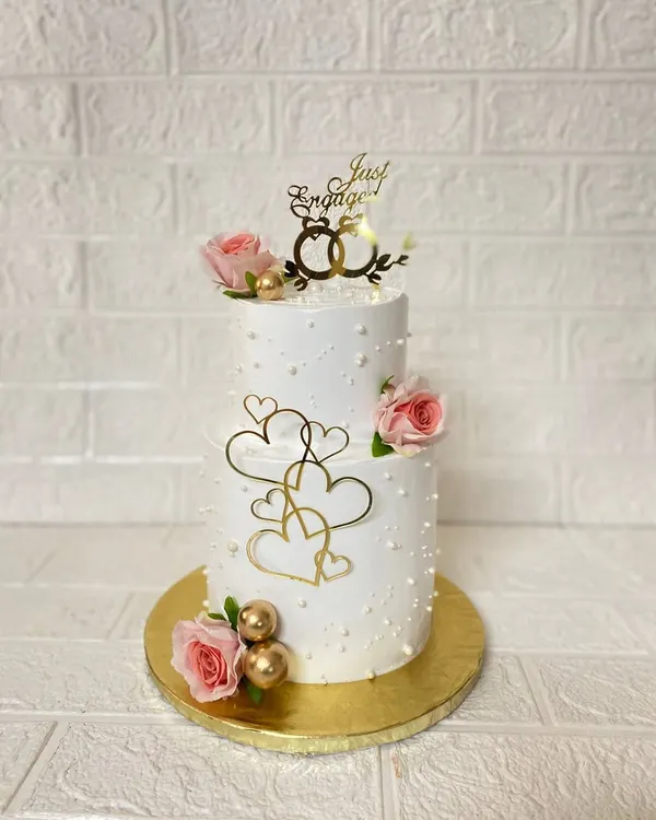 A beautiful two-tier vanilla cake we curated for an engagement ceremony. It's adorned with fresh roses, pearl details, and elegant gold heart accents.