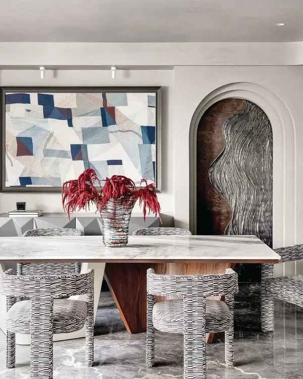 This dining space is a study in texture and form. The woven chairs, marble tabletop, and abstract art create a sophisticated yet inviting atmosphere. The arched doorway reveals a custom art panel, adding another layer of craftsmanship to the room.