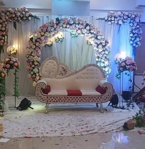 A classic engagement stage setup featuring an ornate white diwan sofa for the couple. The backdrop is a full circular arch made of dense pastel pink and white roses, complemented by matching floral stands and soft lighting.