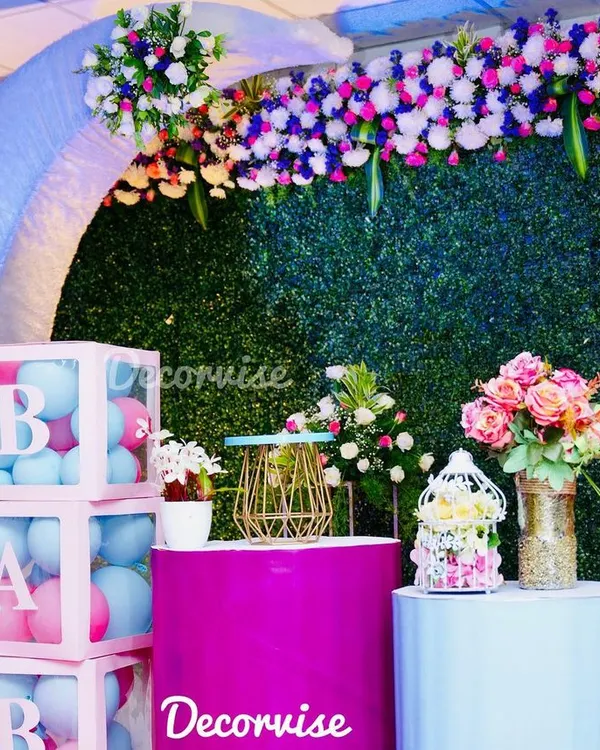 This modern baby shower decor combines several popular elements. It features a lush artificial green wall, a pastel blue arch, and clear acrylic boxes spelling "BABY" filled with pink and blue balloons for a gender-neutral theme.