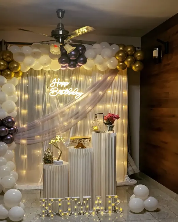 An elegant birthday party setup at home, featuring a white and gold balloon arch, a sheer curtain backdrop with fairy lights, and illuminated marquee letters spelling out the name 'KUMAR'.