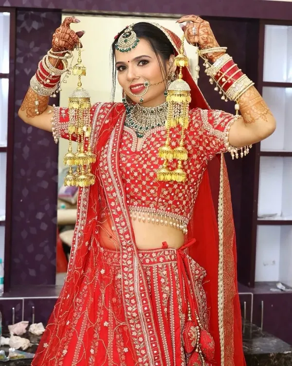 A joyful bride ready for her ceremony. This look features a classic red lip, defined eyes, and a flawless base, perfectly complementing her red lehenga and green Kundan jewellery.