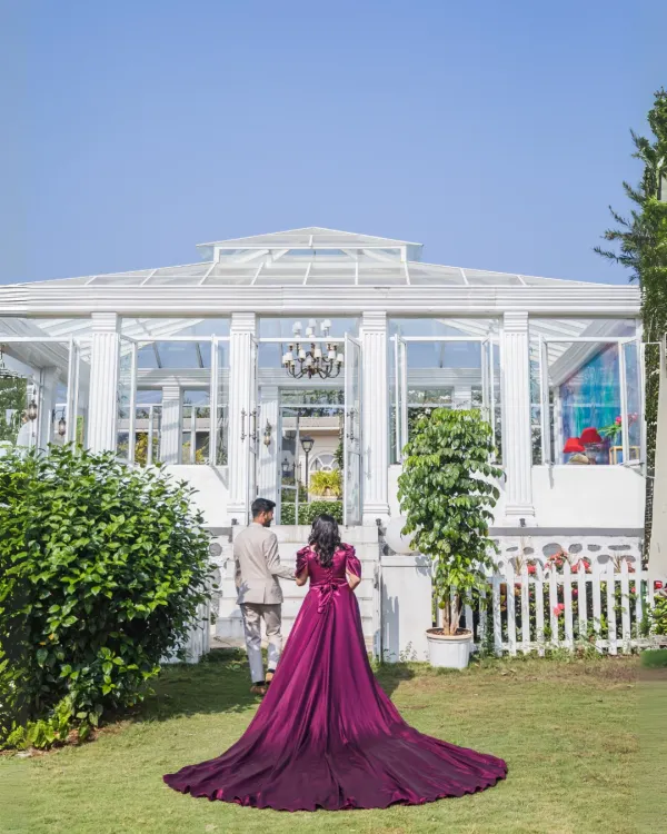 A dramatic and elegant shot from behind, capturing the bride's magnificent flowing gown as the couple walks towards a beautiful white conservatory. A perfect blend of fashion and romance.