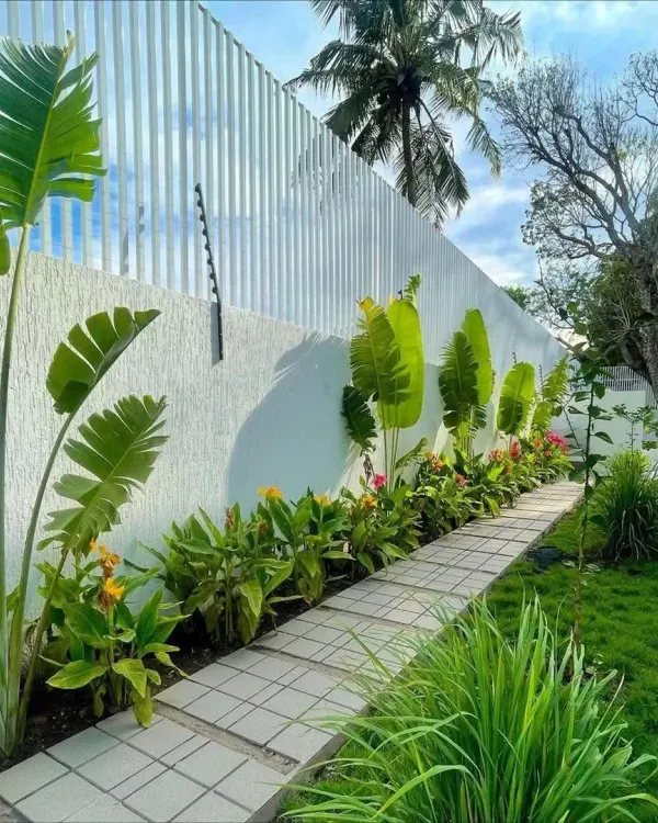 A clean, modern pathway made of concrete pavers, flanked by a narrow planting bed with tropical plants. The design is simple, elegant, and guides you along the property wall.