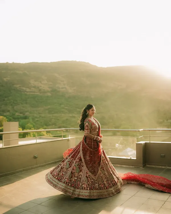 A bride twirls in her magnificent red lehenga on a rooftop at sunset, creating a breathtaking and epic portrait against the mountain landscape.